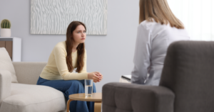 | Nashville Mental Health Woman in a yellow sweater sits on a beige sofa, talking with a therapist at a small table with a glass of water nearby, in a bright room.
