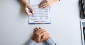 | Nashville Mental Health Two people at a desk: one writes on a document on a clipboard while the other rests their hands folded on the table.