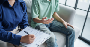 Person in blue writes on a clipboard while another in green sits on a sofa and speaks with a hand gesture.