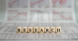 Wooden letter blocks spell RESEARCH on a newspaper with blurred stock charts in the background.