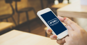 Smartphone screen showing '#FOMO' with 'Fear of Missing Out' in a cafe setting.