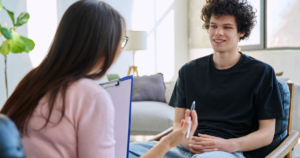 Therapist seated with clipboard talking to a smiling young man in a bright therapy room, fostering conversation.