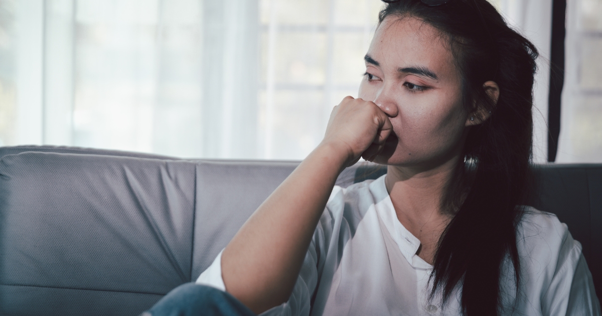 Woman seated on a sofa with her hand covering her mouth, looking thoughtful or worried in a casual setting.