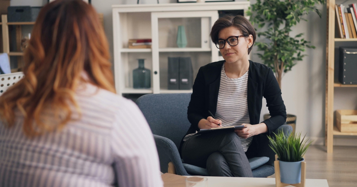 | Nashville Mental Health Two women sit facing each other in a modern office; one with short dark hair and glasses takes notes on a clipboard while the other, seen from the back, listens across the table.
