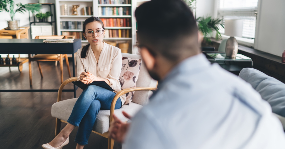 Therapist seated with notes, listening to a client in a cozy living-room therapy space.
