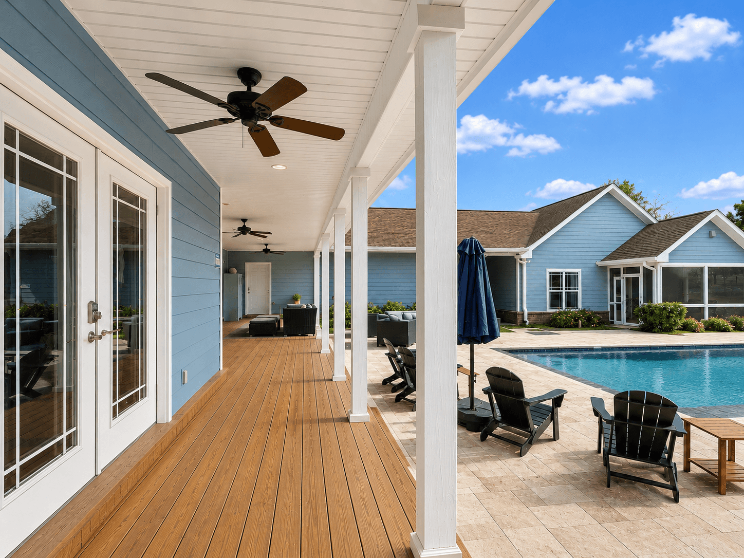 Covered wooden deck along a blue building with white columns, ceiling fans, and outdoor seating by a pool area