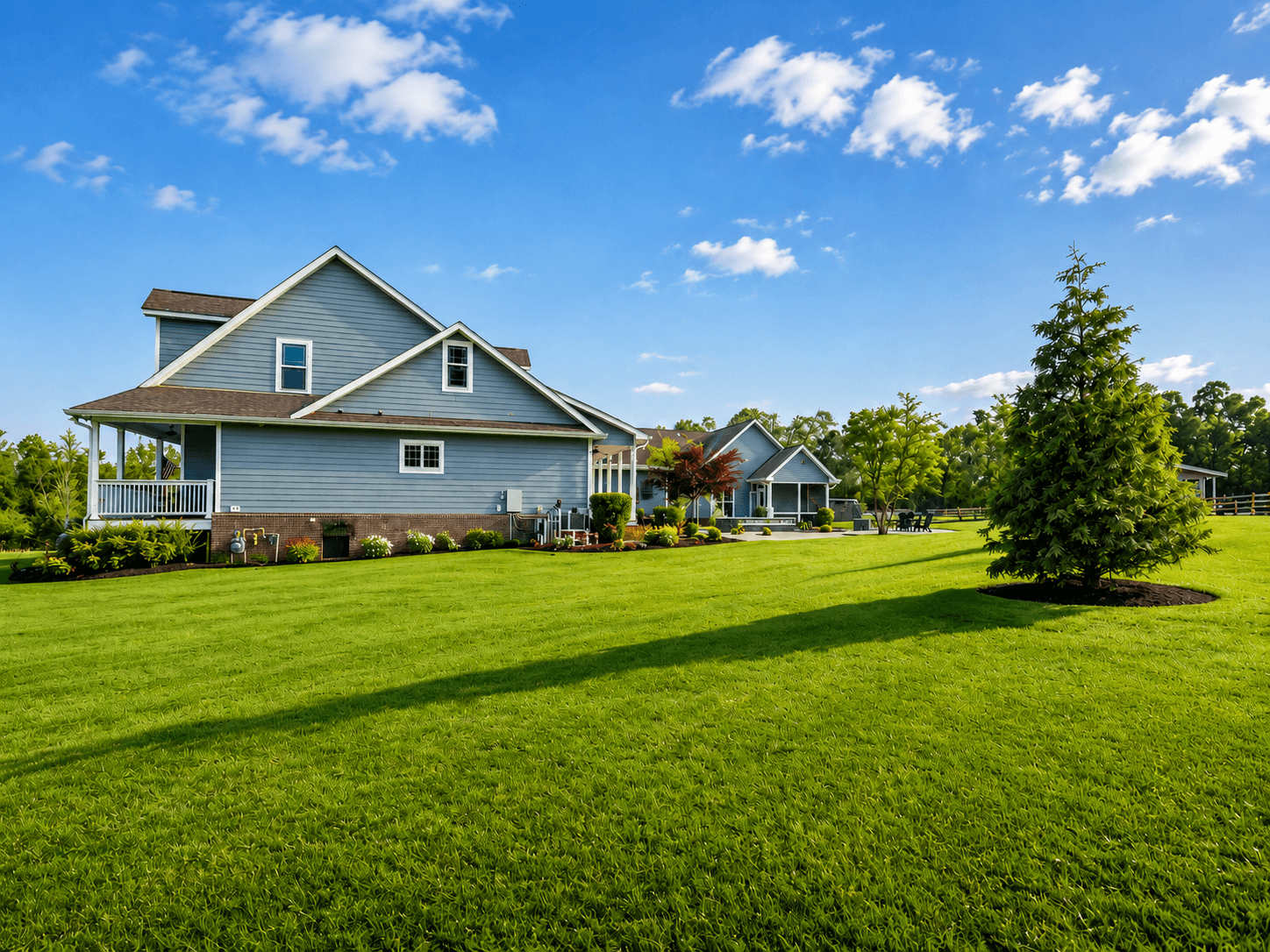 Blue two-story house with white trim, wraparound porch, and a lush green lawn under a bright blue sky.