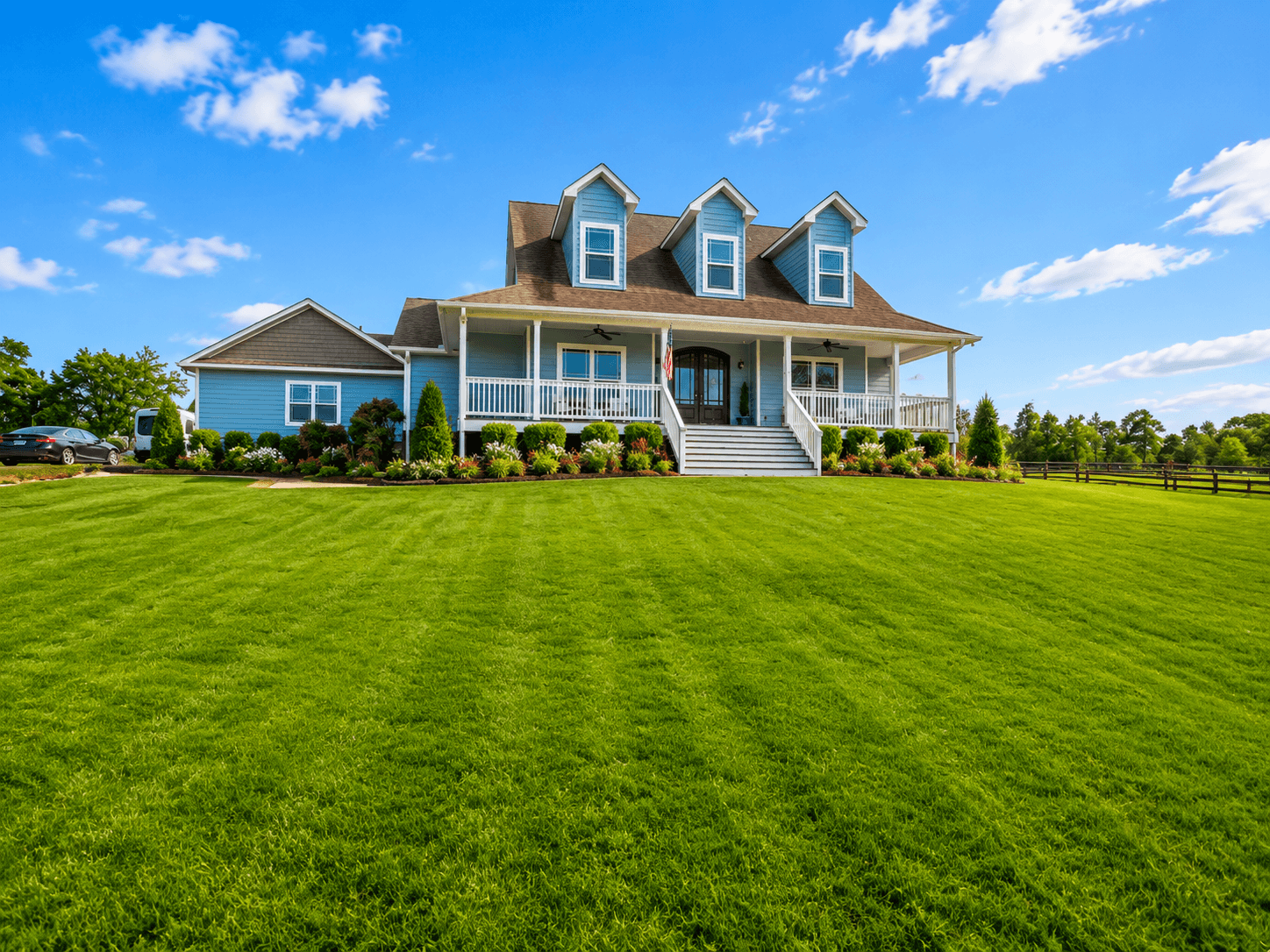 Blue two-story house with three dormer windows, white front porch railings, and a manicured lawn.