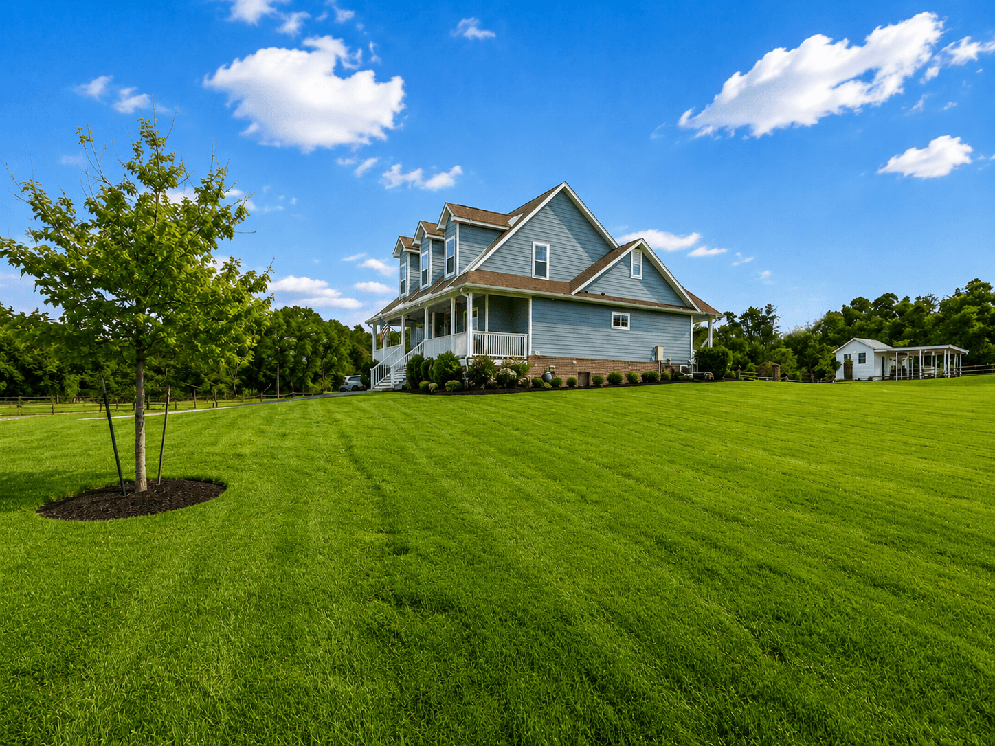 Blue two-story house with a wraparound porch on a manicured green lawn under a bright blue sky.
