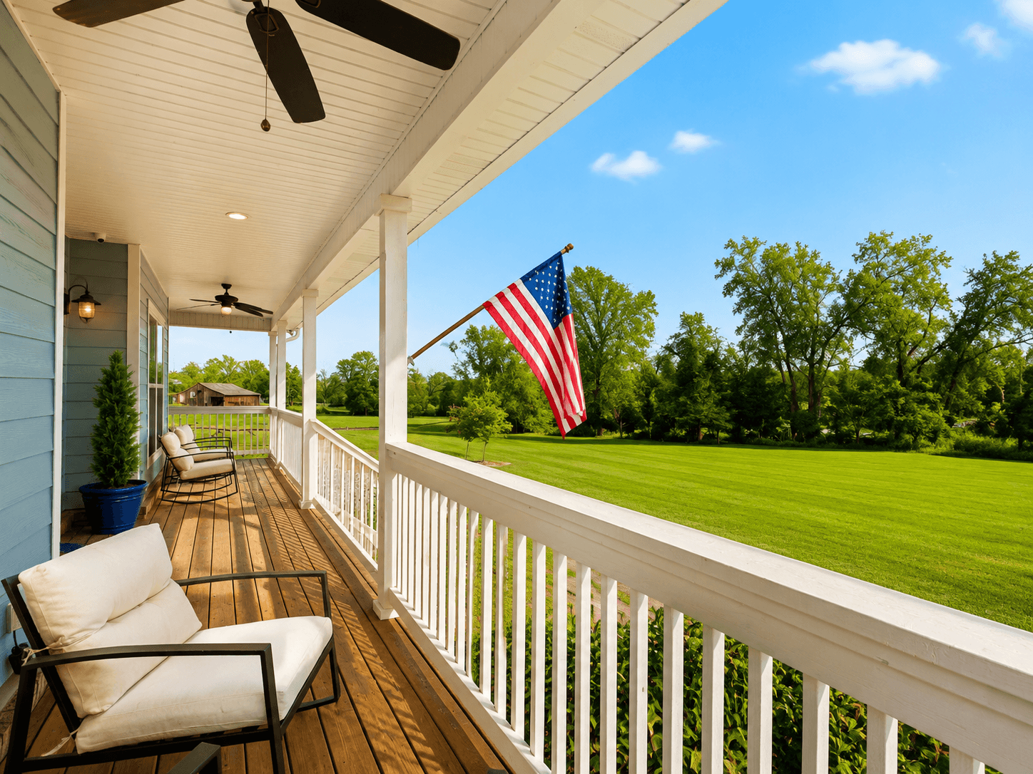 Front porch of a blue house with white railing, beige outdoor chairs, and an American flag on a pole over a green lawn.
