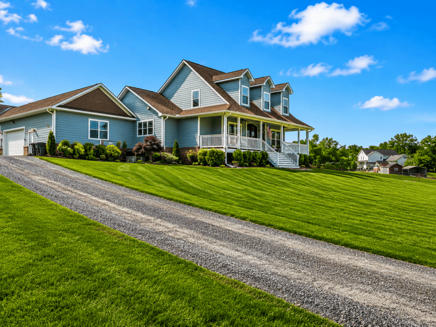 Blue two-story house with a wraparound porch on a sunny day, green lawn, and a gravel driveway in the foreground.