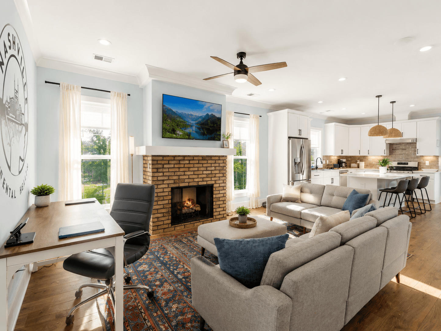 Bright open-concept living room with a brick fireplace, wall-mounted TV, and a connected white-kitchen layout.
