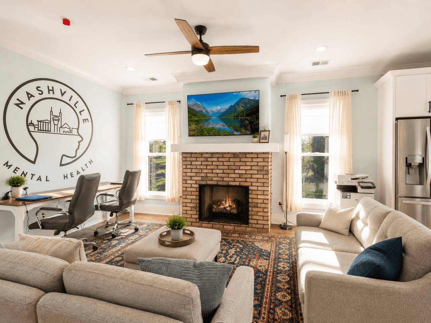Office-like living room with a brick fireplace, TV above it, and a large Nashville Mental Health logo on the left wall; neutral sofas, rug, windows with sheer curtains, and a ceiling fan.