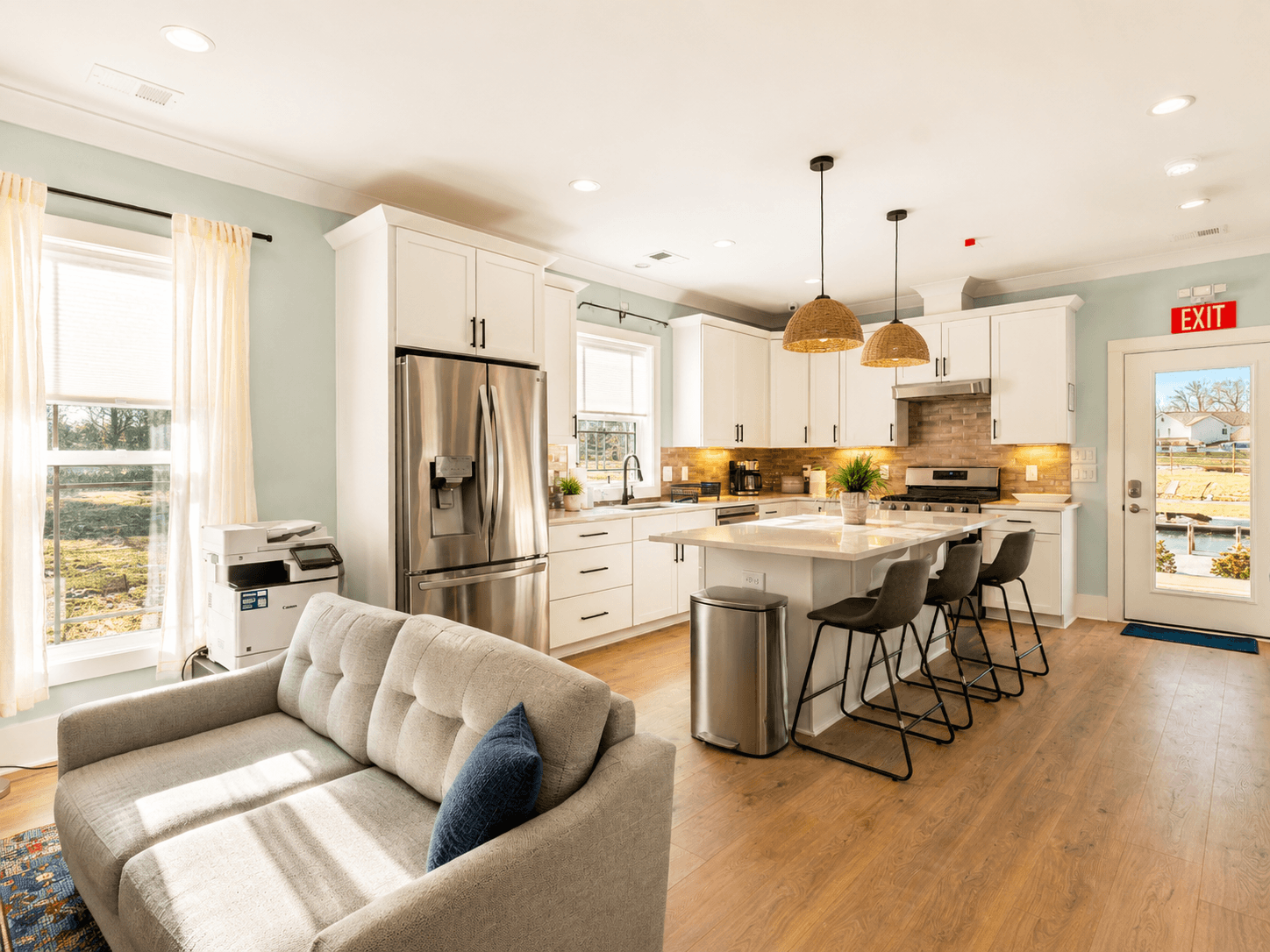 Open-plan kitchen and living area with white cabinets, stainless steel fridge, island seating, and wicker pendant lights. Similar neutral tones and daylight fill the space.