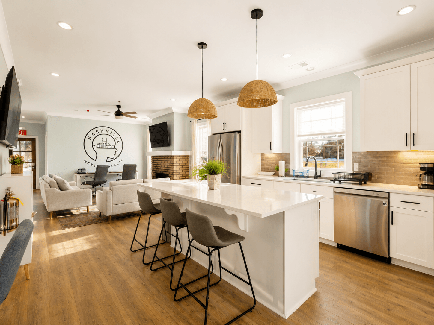 Open-concept kitchen and living area featuring a white island with four gray bar stools, wicker pendant lights, stainless steel appliances, and beige seating area; wall art with a circular logo in the background.