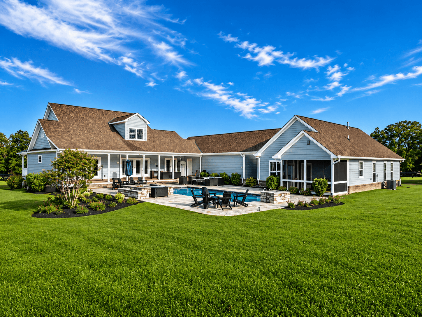 Wide view of a blue house with a brown roof, a screened porch, and a rectangular pool surrounded by a stone patio and outdoor seating.