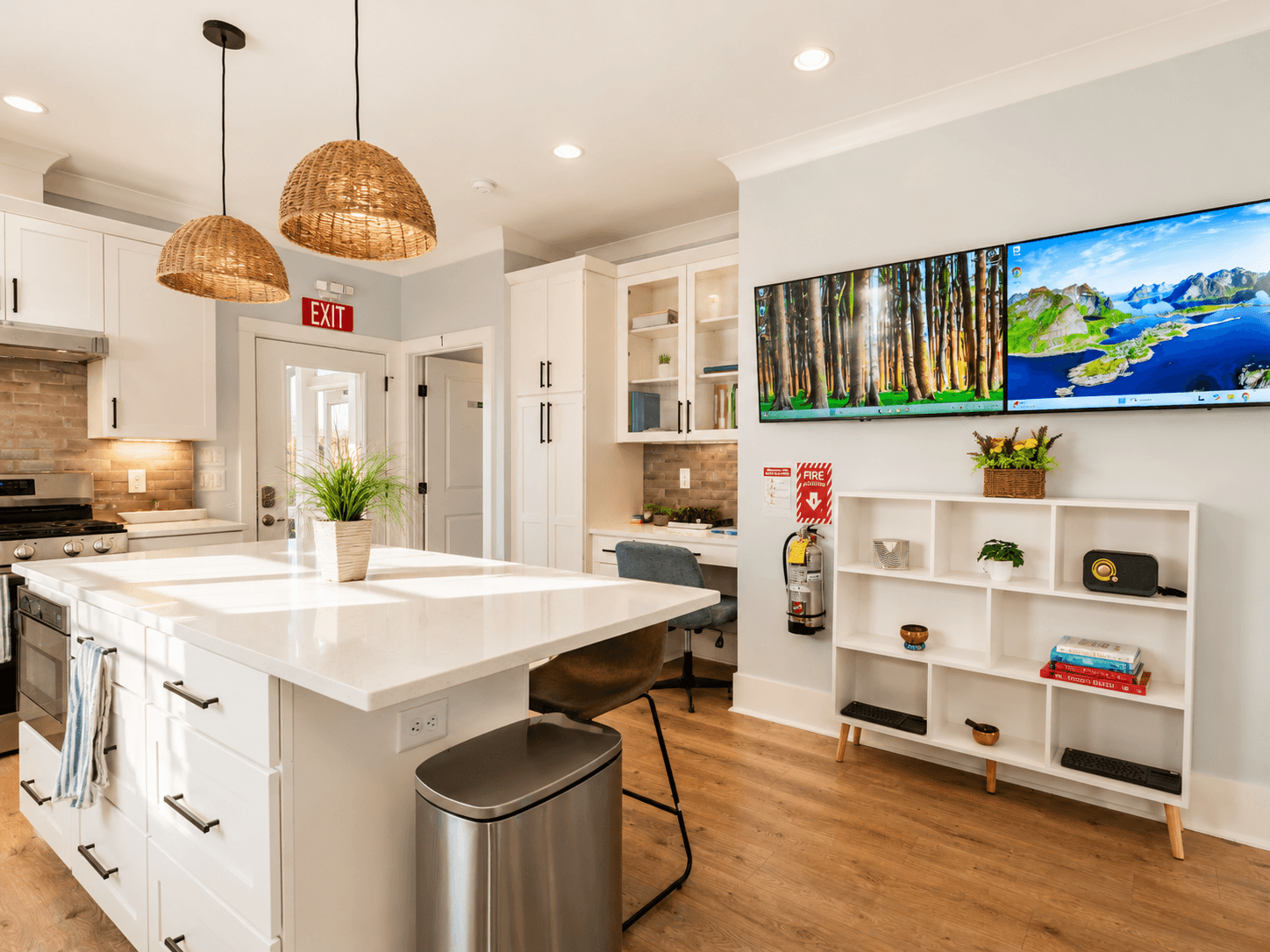 Open-concept kitchen with white cabinets and a large island, wicker pendant lights, a desk nook, exit sign, and a wall-mounted dual-monitor TV above a white cubby shelf.