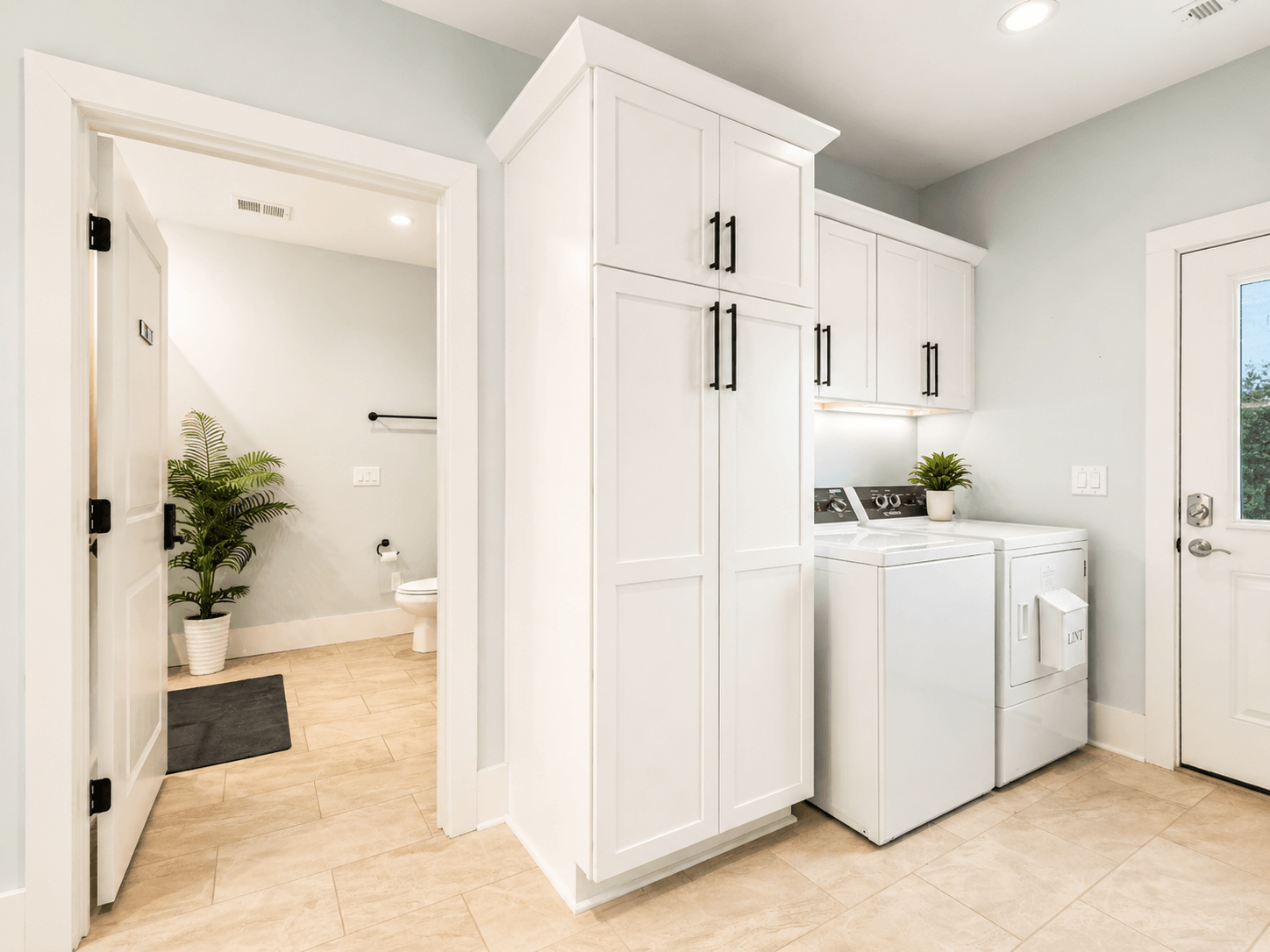 Bright laundry room with tall white cabinets, a washer and dryer, and a small plant; doorway to a bathroom on the left.
