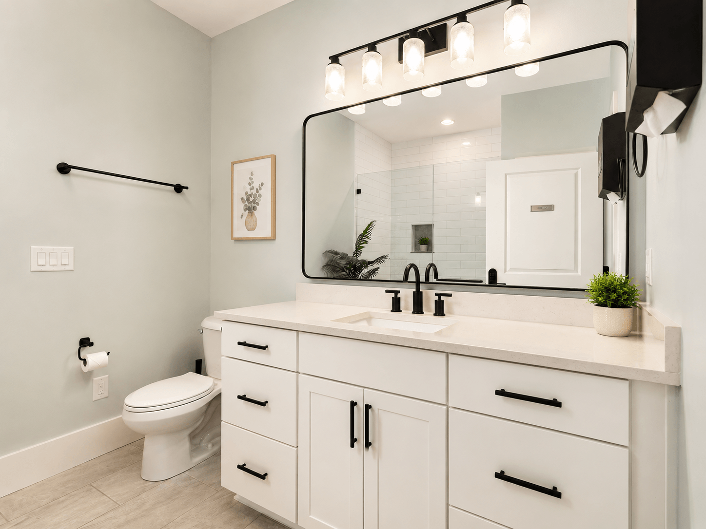 Bright, modern bathroom with white vanity, black handles, and a large framed mirror; four-light fixture above and a plant on the counter.