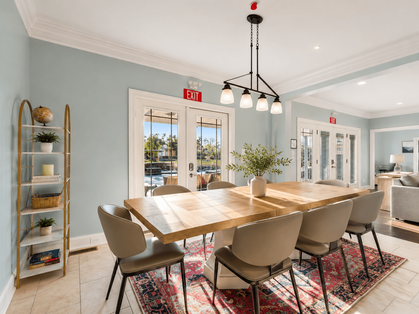 Bright dining room with a large wooden table, six gray chairs, and a vase of greenery under a modern light fixture, near glass doors to the outdoors.