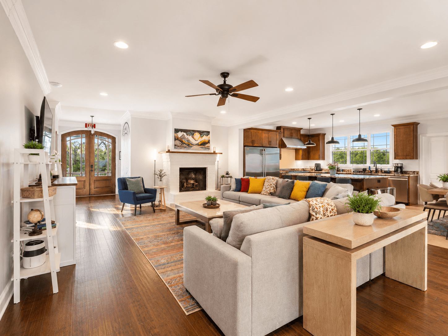 Bright open-concept living room and kitchen: gray sectional with colorful pillows facing a white fireplace, hardwood floors, and a ceiling fan, with a wooden door nearby.