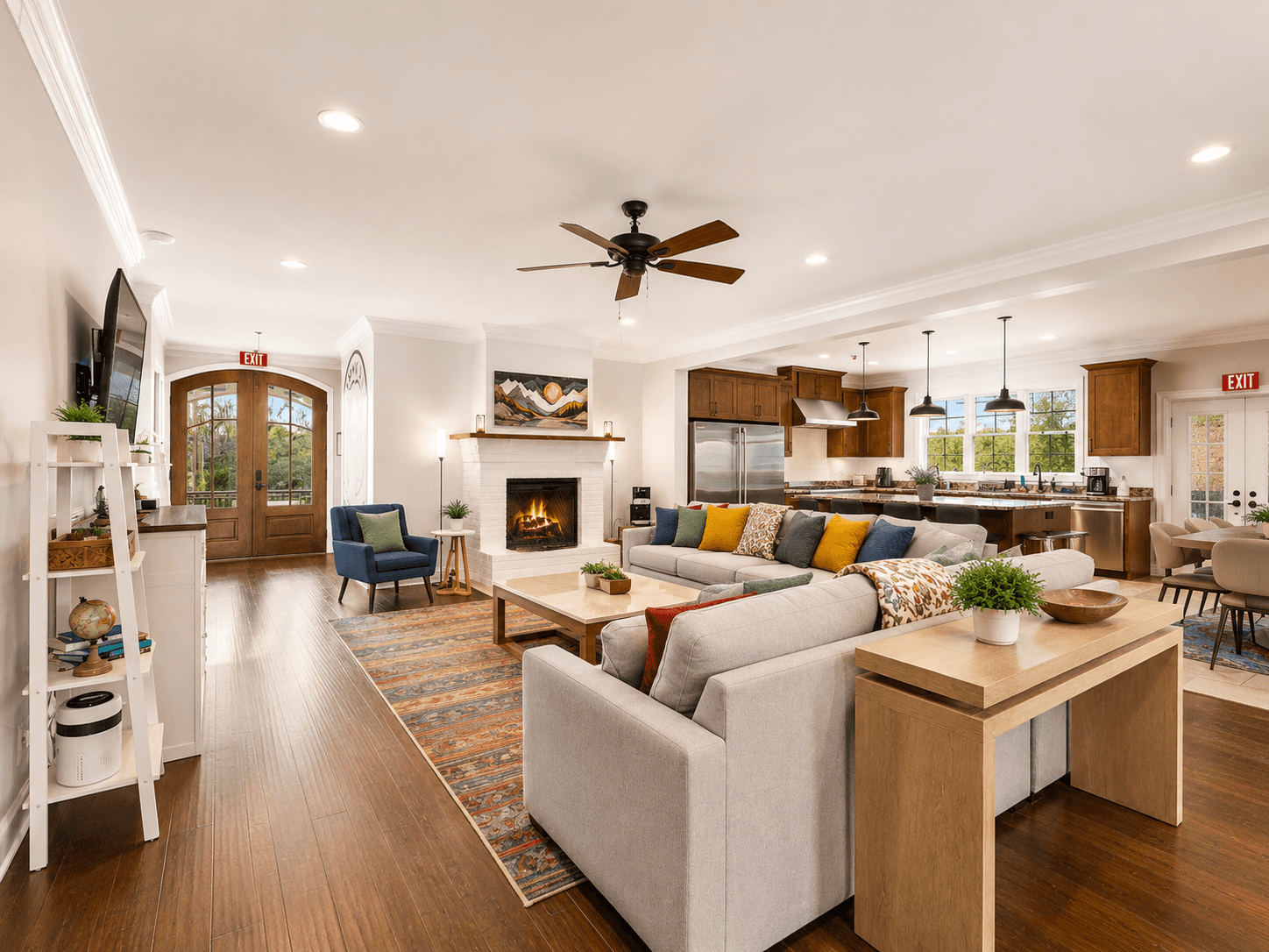 Spacious open-plan living room with gray sectional, colorful cushions, and a lit fireplace adjacent to a kitchen island and wooden cabinets.