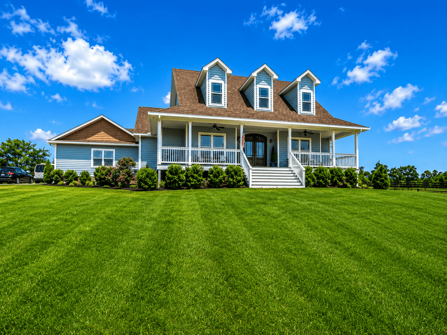 Blue two-story house with three dormer windows, wraparound porch, American flag, and a well-kept green lawn under a bright blue sky.