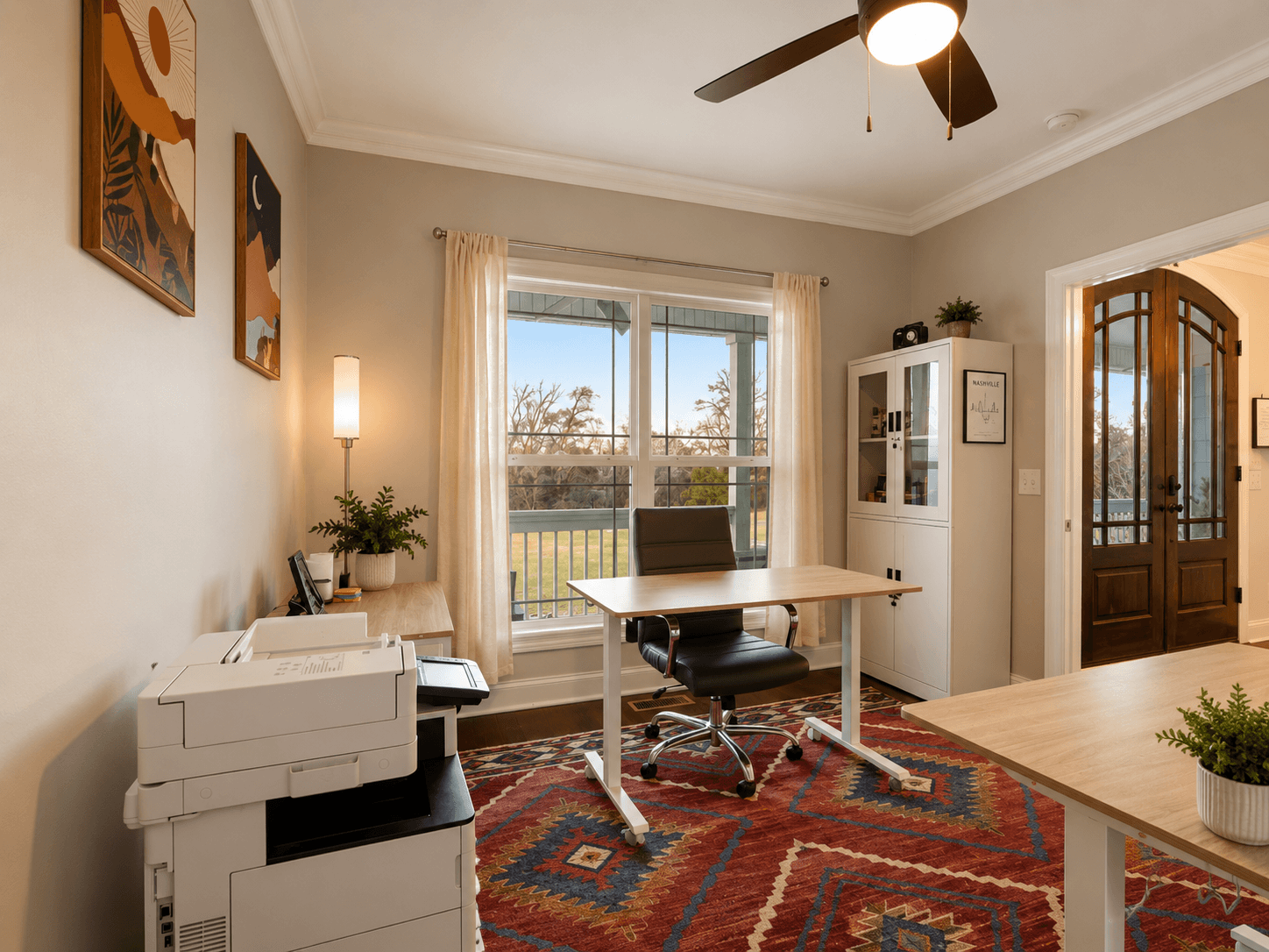 Home office with a wooden desk and black leather chair by a large window; printer on a side table and a colorful rug beneath.