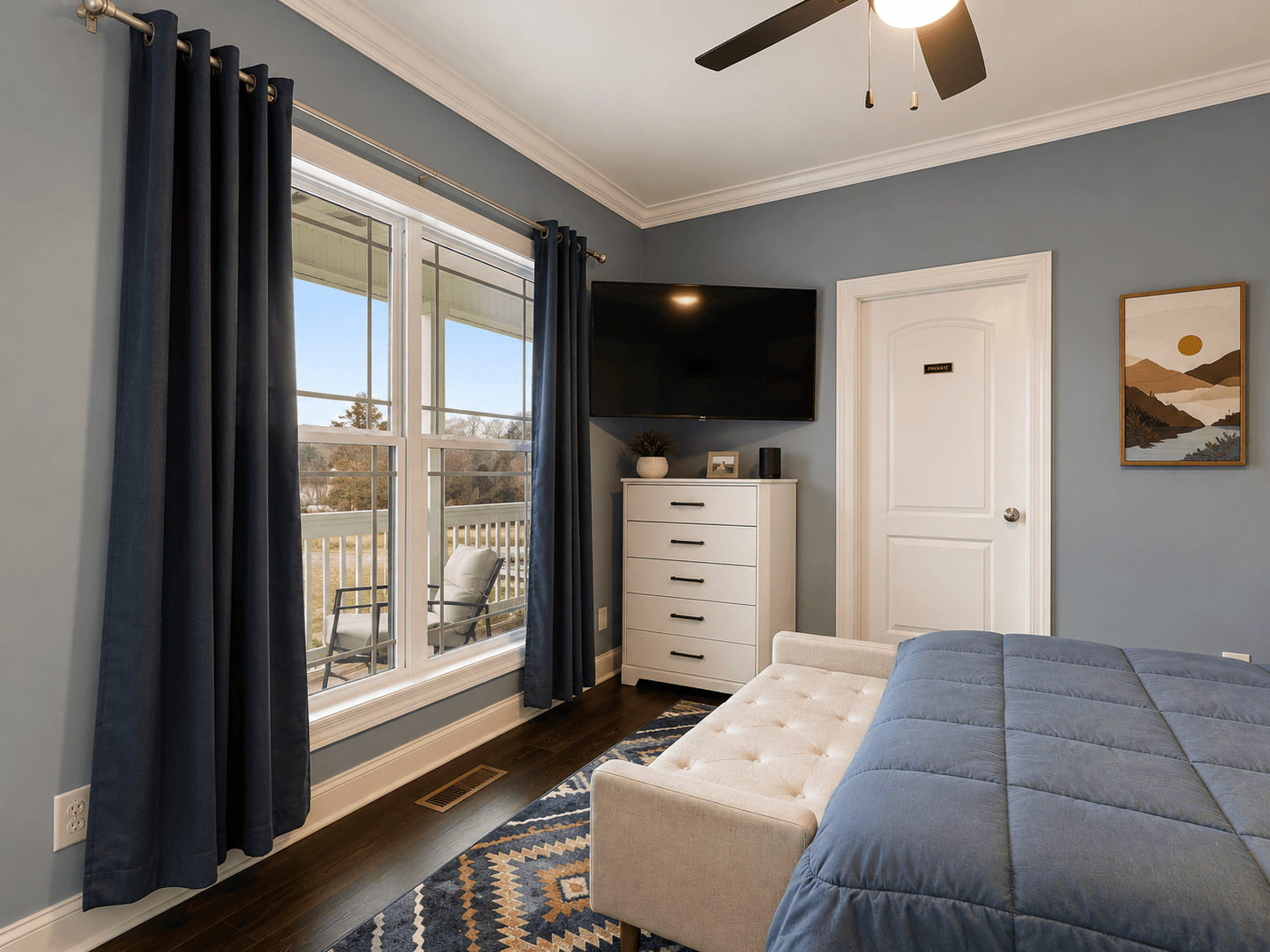 Bedroom interior with blue walls, large window with navy curtains, white dresser, wall-mounted TV, and a bed with a blue quilt.