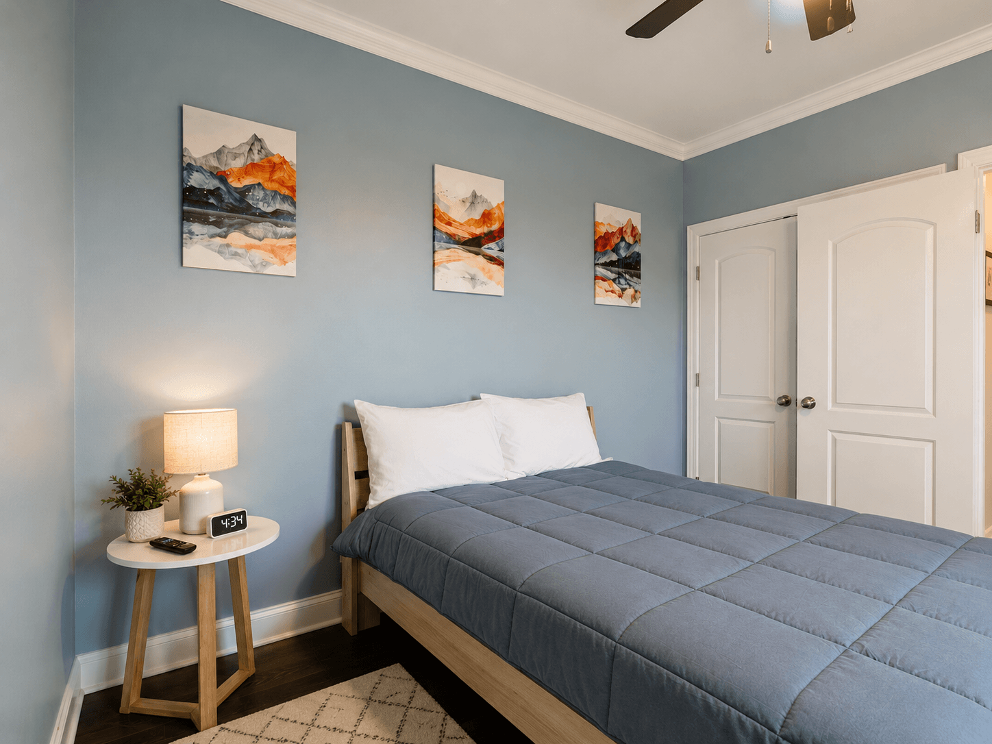Bedroom with blue walls, wooden bed frame, blue quilt, and white pillows; three mountain artworks above the bed and a nightstand with lamp and plant nearby.
