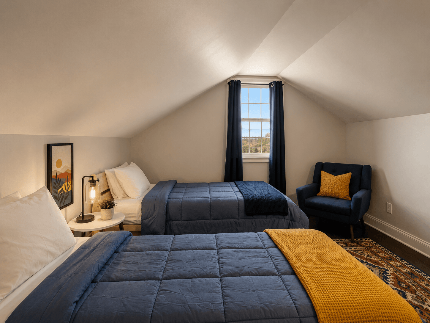 Attic bedroom with two twin beds in blue quilts, a navy window curtain, and a blue armchair with a yellow cushion.