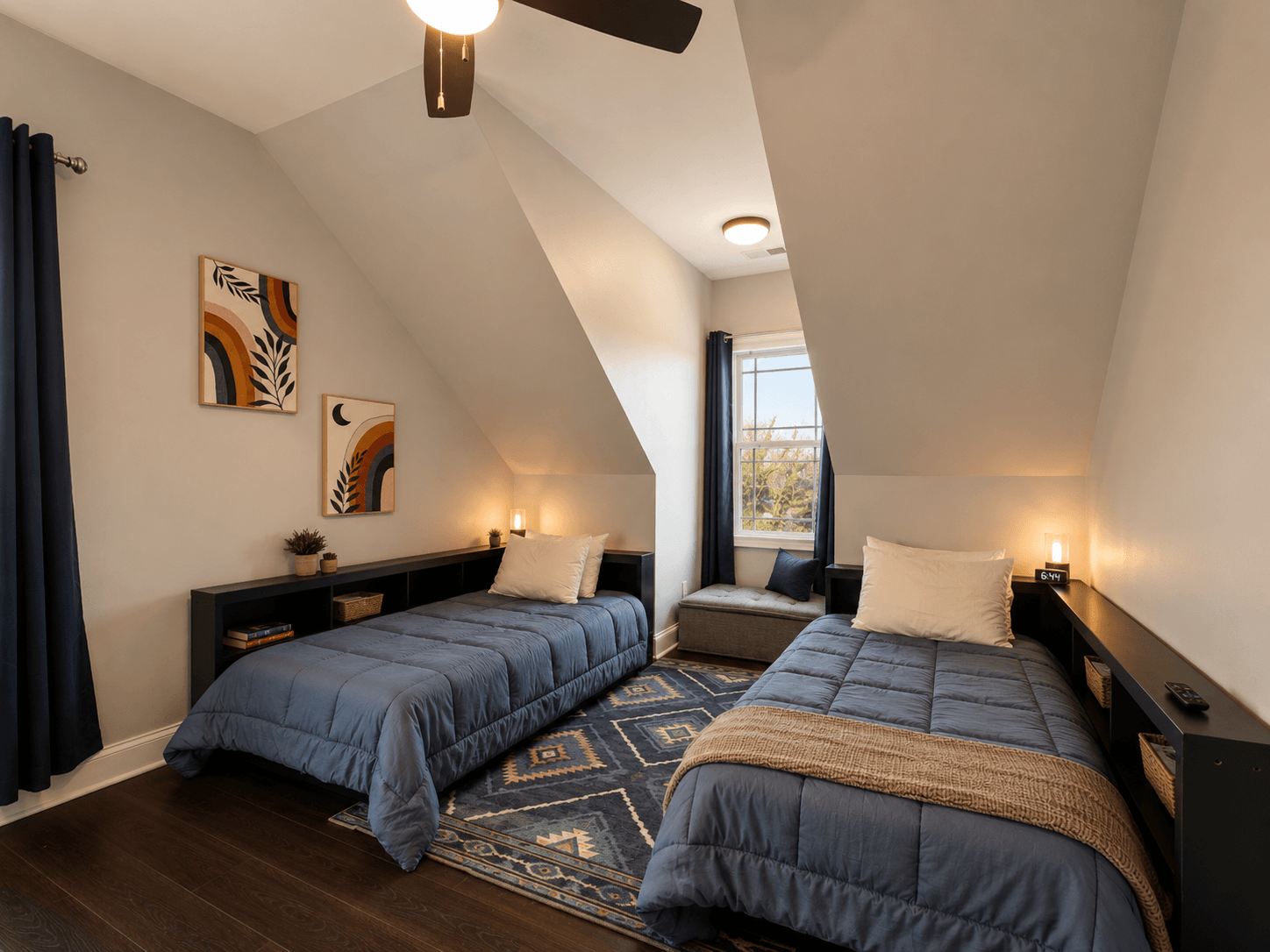 Two twin beds with blue quilts in an attic bedroom, featuring a window seat and navy curtains.