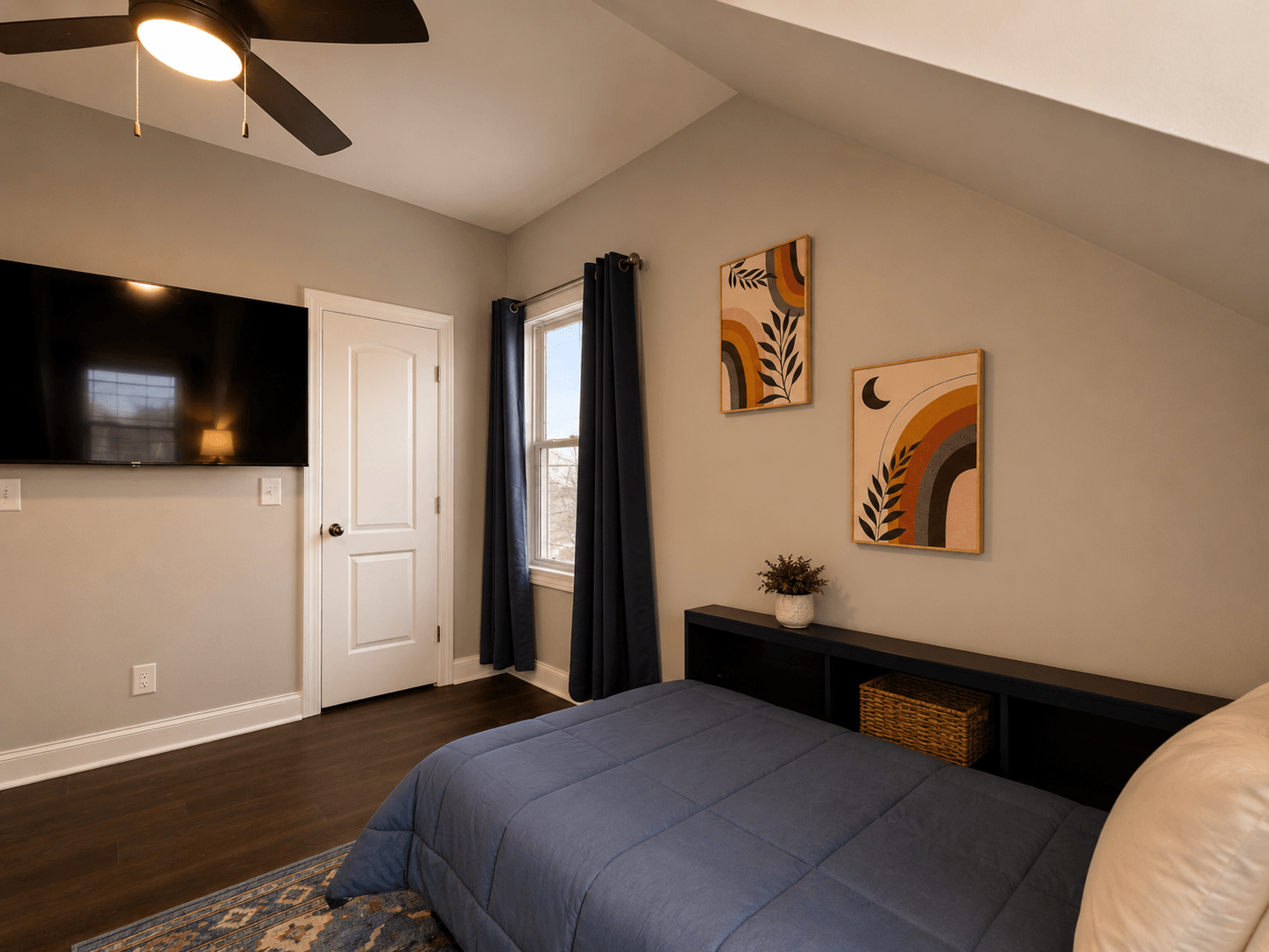 Bedroom with a blue quilted bed, a wall-mounted TV, white door, and dark curtains by a window; decorative art on the wall and a low black shelf with a woven basket.