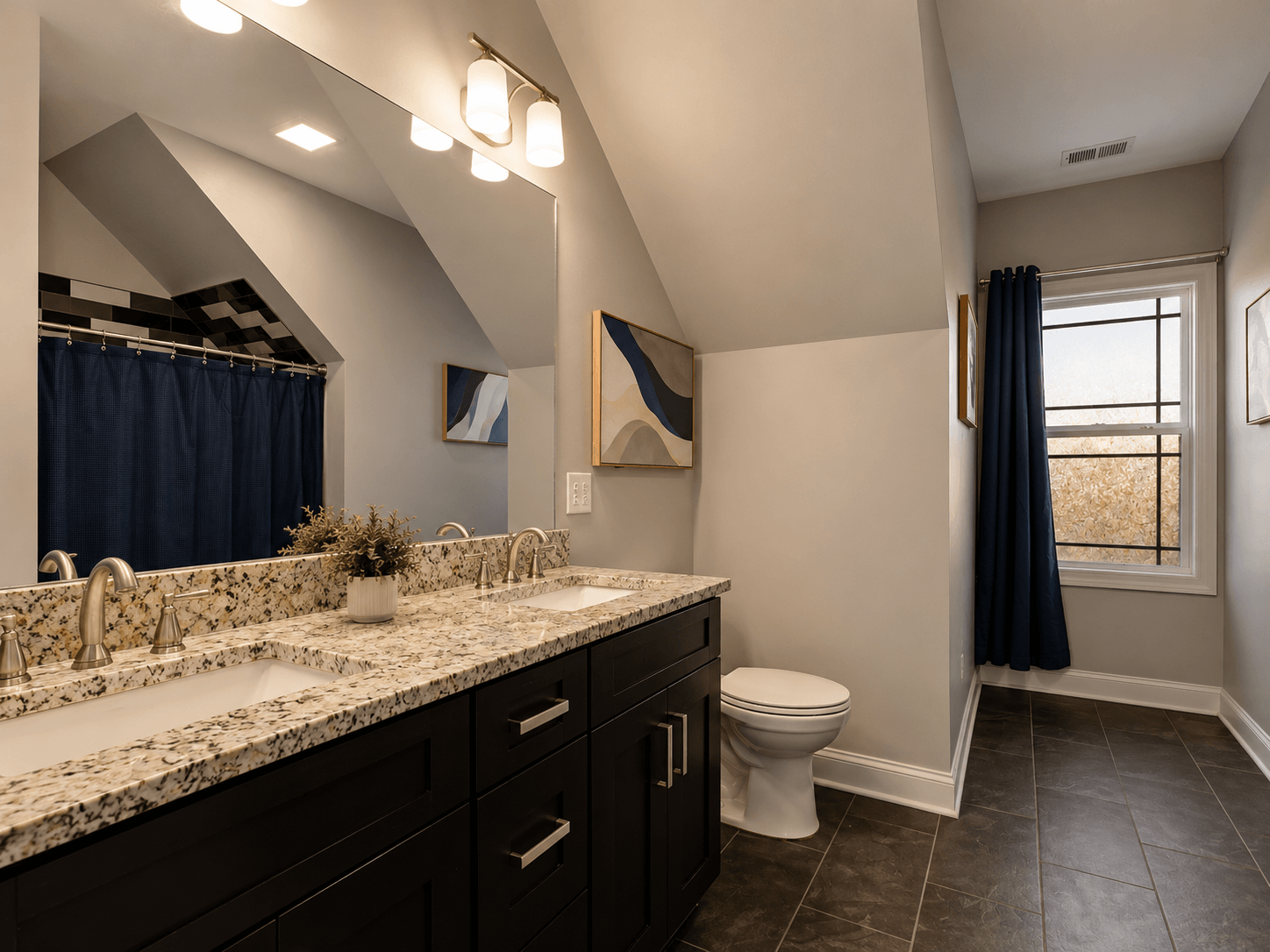 Wide view of a modern bathroom showing a granite double vanity with two sinks, dark cabinets, and a large mirror with warm lighting above.