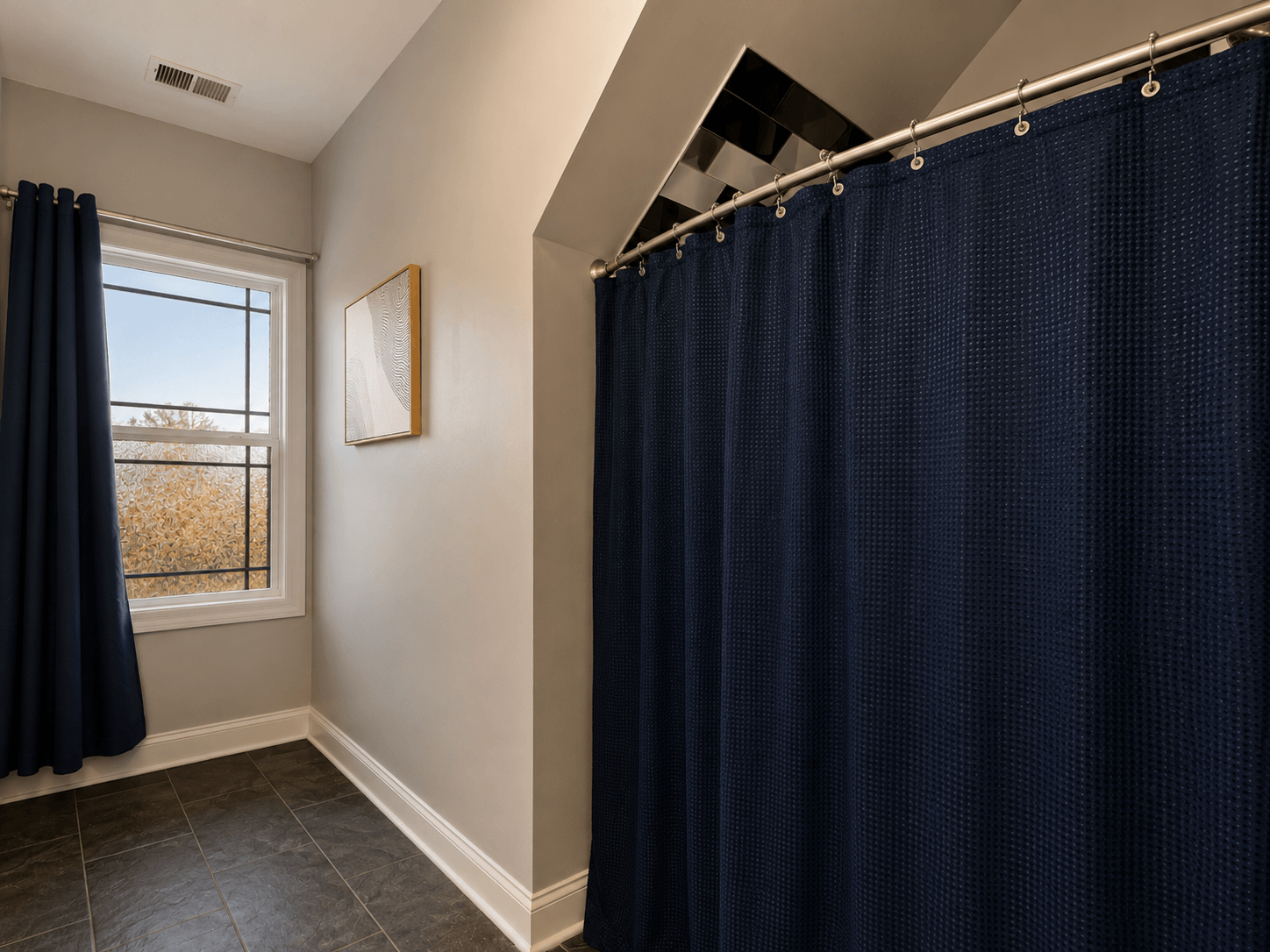Dark blue shower curtain along a tiled bathroom with a window and blue drapes, beige walls, and a framed artwork on the wall.