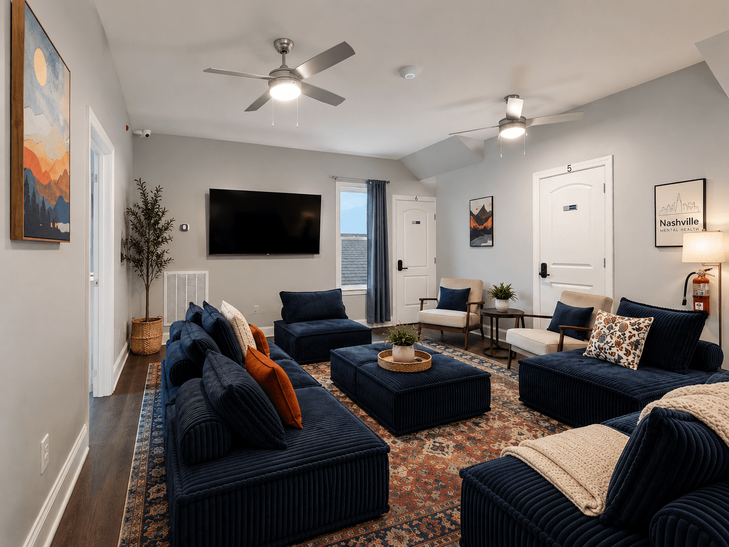 Cozy living room with navy corduroy sofas, central tufted ottoman, and a wall-mounted TV on light gray walls.