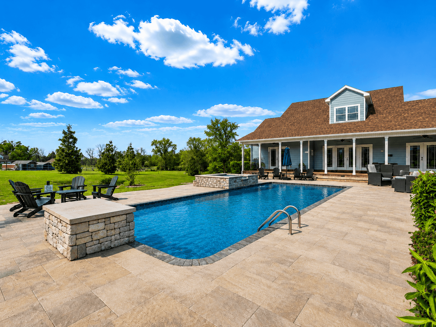 Residential backyard pool with stone planter borders, beige paver deck, and a blue house with a covered patio behind.