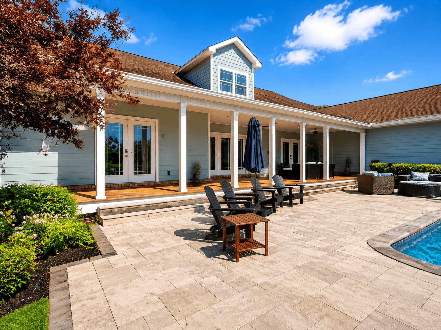 Backyard of a blue house with a covered porch, white columns, patio chairs, and a pool on the right under a bright sky.