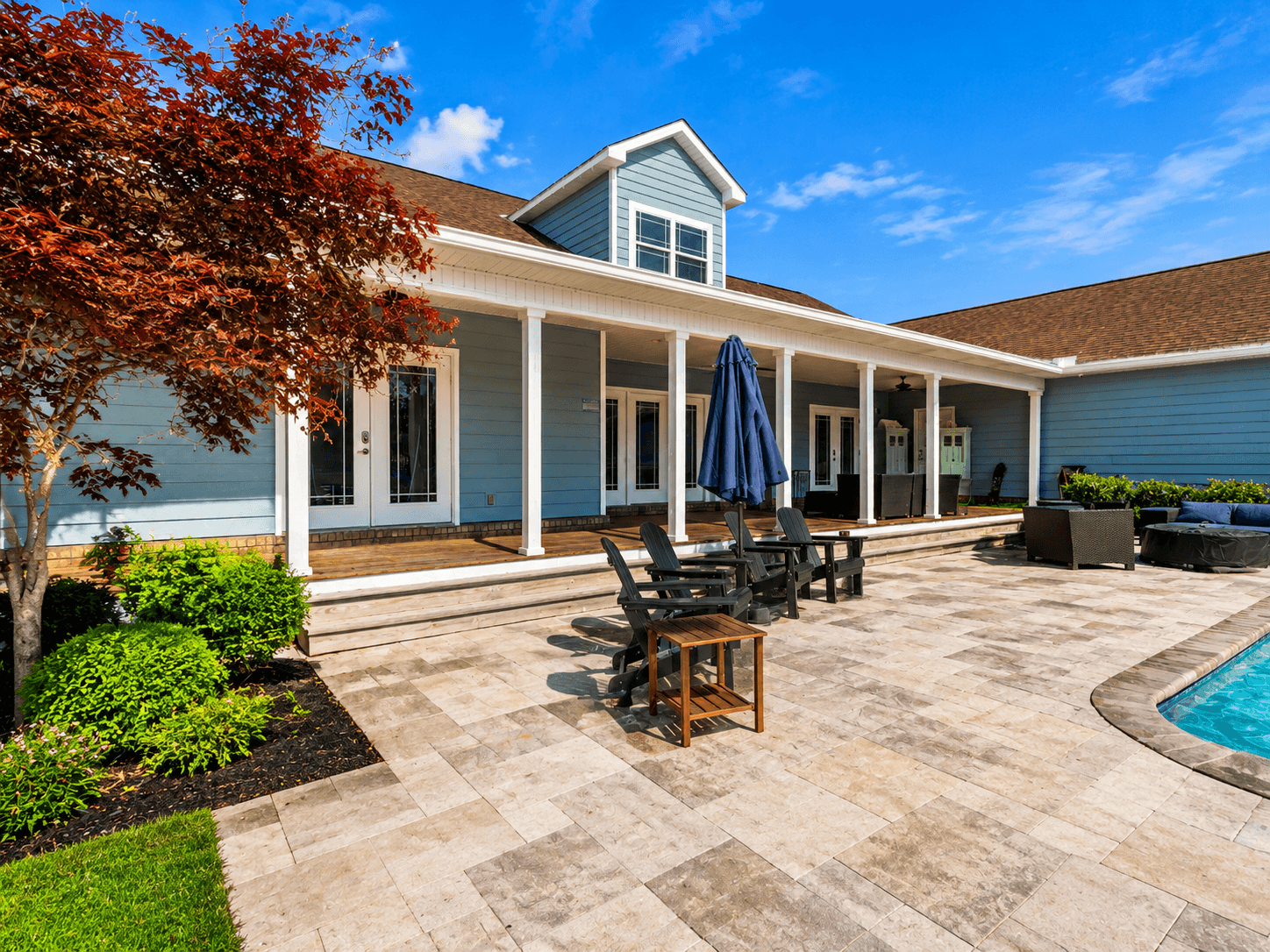Backyard patio of a blue house with white trim, stone deck, and poolside seating under a bright sky.